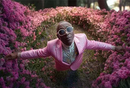 A man in a pink sequined suit and sunglasses floats face-up over a field of pink flowers